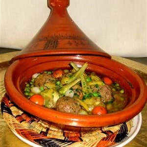 May include: A terracotta tagine with a conical lid containing a stew of meatballs, green peas, sliced artichokes, and cherry tomatoes. The cooking vessel features a wide, shallow base and a tall, pointed top with dark etched decorative patterns.