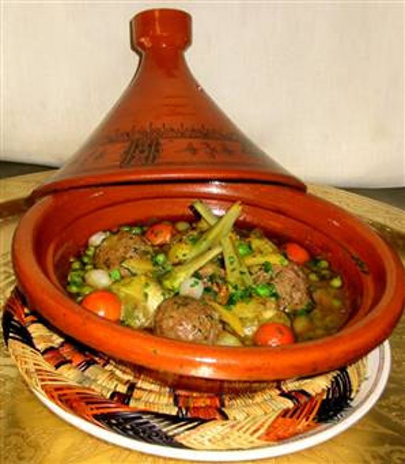 May include: A terracotta tagine with a conical lid containing a stew of meatballs, green peas, sliced artichokes, and cherry tomatoes. The cooking vessel features a wide, shallow base and a tall, pointed top with dark etched decorative patterns.