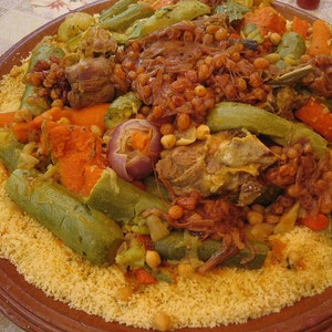 May include: A large round terracotta platter filled with a mound of yellow couscous topped with cooked meat, chickpeas, sliced carrots, green zucchini, and caramelized onions. The dish is arranged in a circular pattern on the plate.