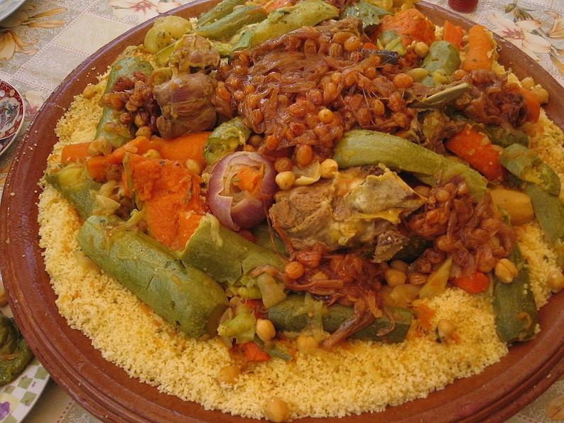 May include: A large round terracotta platter filled with a mound of yellow couscous topped with cooked meat, chickpeas, sliced carrots, green zucchini, and caramelized onions. The dish is arranged in a circular pattern on the plate.