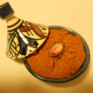 May include: A ceramic tagine with a conical lid featuring blue, red, and black hand-painted geometric patterns. The base is filled with a coarse, orange-brown spice blend and topped with a single dried pink rosebud in the center.