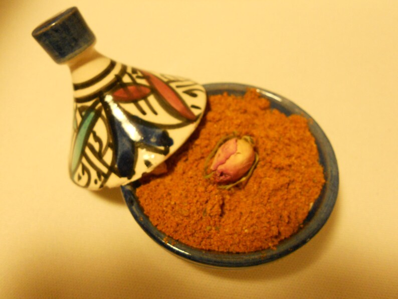 May include: A ceramic tagine with a conical lid featuring blue, red, and black hand-painted geometric patterns. The base is filled with a coarse, orange-brown spice blend and topped with a single dried pink rosebud in the center.