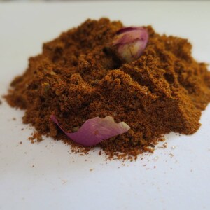 May include: A mound of finely ground, deep reddish-brown spice blend with a coarse, textured appearance. Two dried, light purple rose petals are nestled into the top and side of the spice pile, providing a contrast in color and texture.