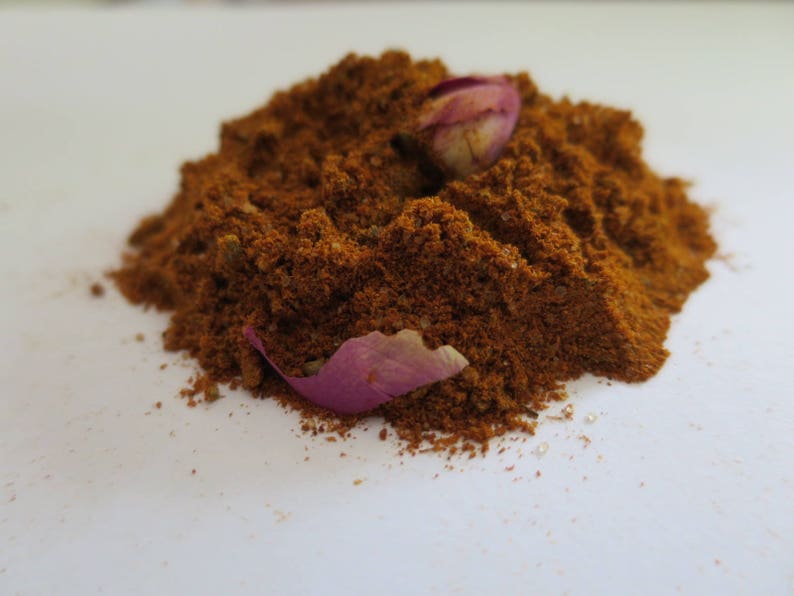 May include: A mound of finely ground, deep reddish-brown spice blend with a coarse, textured appearance. Two dried, light purple rose petals are nestled into the top and side of the spice pile, providing a contrast in color and texture.