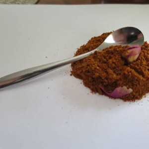 May include: A polished silver-toned metal spoon resting on a mound of finely ground, rust-colored spice powder. Two small, dried pink rose petals are partially embedded within the textured pile of spice, which sits on a plain white surface.