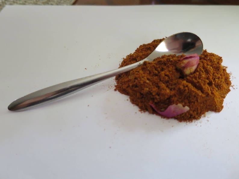 May include: A polished silver-toned metal spoon resting on a mound of finely ground, rust-colored spice powder. Two small, dried pink rose petals are partially embedded within the textured pile of spice, which sits on a plain white surface.