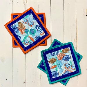 May include: Two square quilted pot holders with a marine life pattern. The fabric features fish, coral, and seahorses in shades of blue, orange, and brown on a light blue background. Each pot holder has a blue border and an orange or teal edge.