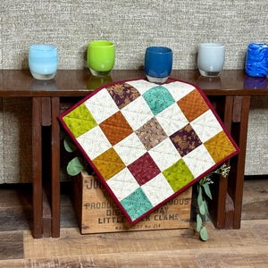 May include: A small, dark brown wooden table displays five colorful glass tumblers and a quilted square. The tumblers are various colors, including blue, green, and gray. The quilt features a patchwork design with squares of different patterns and colors.