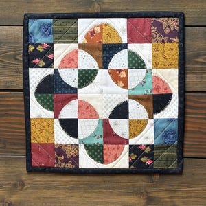 May include: A square quilted wall hanging featuring a patchwork design of various fabric squares in shades of red, blue, green, yellow, and black. The center displays four circular motifs created from curved pieced fabric segments with visible stitching.