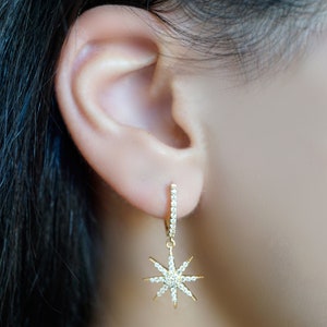 Puede incluir: Pendientes de aro dorados con forma de estrella y piedras de circonita cúbica transparentes. Los pendientes tienen un diseño simple y minimalista.
