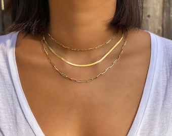 May include: Three gold necklaces layered on a person's neck. The top necklace is a thin chain with small, square links. The middle necklace is a thicker, flat chain. The bottom necklace is a chain with larger, square links.