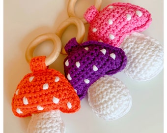 May include: Three crocheted mushroom toys with wooden rings. The mushrooms are orange, purple, and pink with white polka dots. The stems are white.