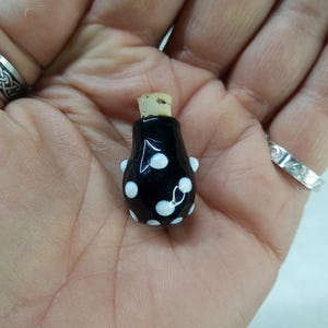 Puede incluir: Una pequeña botella de vidrio negro con un tapón de corcho y lunares blancos.