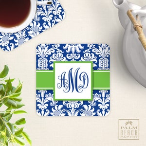 Puede incluir: Un posavasos azul y blanco con estampado de damasco, una banda verde y un cuadrado blanco con el monograma "AMD" en negro. El posavasos est&aacute; sobre una superficie blanca.