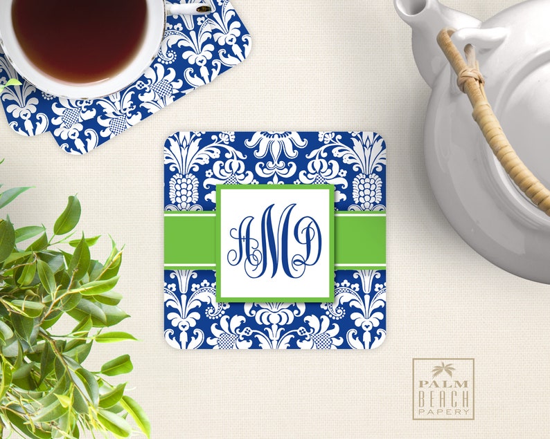 Puede incluir: Un posavasos azul y blanco con estampado de damasco, una banda verde y un cuadrado blanco con el monograma "AMD" en negro. El posavasos est&aacute; sobre una superficie blanca.