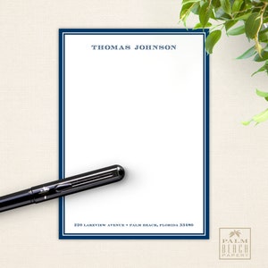May include: A white notepad with a blue border and the text "THOMAS JOHNSON" at the top. The notepad has the address "220 LAKEVIEW AVENUE * PALM BEACH, FLORIDA 33480" printed at the bottom. The notepad is on a beige surface with a black pen lying on top of it.