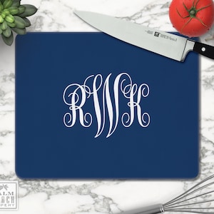 May include: A navy blue cutting board with white monogram initials. A stainless steel chef's knife, tomato, and garlic are on the board. A whisk and a succulent plant are also in the image. The cutting board is a kitchen item.