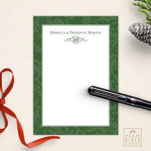 May include: A personalized stationery set featuring a white writing surface with a green floral border. The text "Rebecca & Donovan Martin" is printed at the top, with a decorative flourish. A black pen rests on the stationery, with a red ribbon and pinecone in the background.
