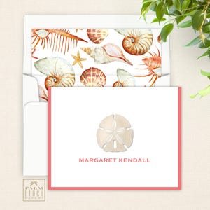 May include: A white card with a pink border and a sand dollar design. The text "MARGARET KENDALL" is printed in black below the sand dollar. The card is in a white envelope with a seashell pattern.