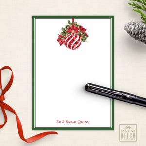 May include: White stationery with a green border and a watercolor Christmas ornament design. The ornament is red and white striped with red poinsettias and greenery. The names "Ed & Sarah Quinn" are printed at the bottom. A black pen and red ribbon are also in the image.
