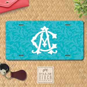 May include: A turquoise blue license plate with a white monogram of the letters "AMC" in a decorative font. The monogram is centered on the plate, which has a floral pattern background. The plate has two holes on the top and two holes on the bottom.