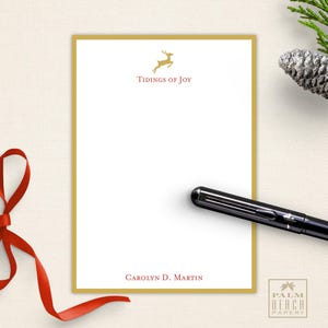 May include: A white stationery set with a gold border, featuring the text "Tidings of Joy" and a gold reindeer illustration. The set includes a black pen and is decorated with a red ribbon. The bottom of the stationery reads "Carolyn D. Martin".