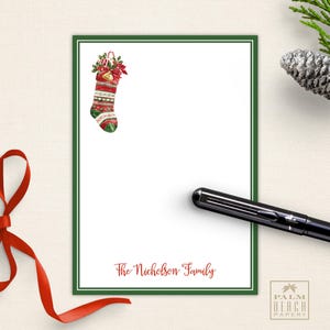 May include: White stationery with a green border and a Christmas stocking illustration. The text "The Nicholson Family" is printed in red. A black pen and red ribbon are also present.