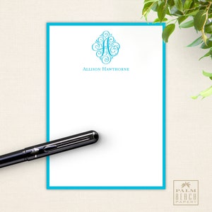 May include: A white notepad with a turquoise border and a turquoise monogrammed initial "A" with a decorative flourish. The notepad has the name "Allison Hawthorne" printed below the monogram. The notepad is on a beige surface.