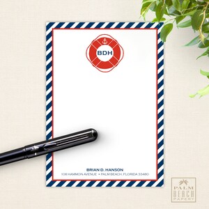 May include: A white notepad with blue and red stripes on the border. The notepad has a red life preserver with a white anchor and the letters "BDH" in the center. The notepad has the text "BRIAN D. HANSON" and "108 HAMMON AVENUE PALM BEACH, FLORIDA 33480" printed at the bottom. The notepad is on a beige surface.