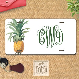 May include: White personalized license plate with a green pineapple illustration on the left side and the monogram "CWD" in green script on the right side. The plate is on a woven straw background.