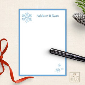 May include: White stationery with a light blue border and snowflake accents. The text "Addison & Ryan" is printed in light blue. A black pen rests on the stationery. A red ribbon and pinecone are in the background.
