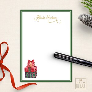 May include: A white notepad with a green border and the name "Alexis Norton" in gold script. A stack of watercolor gift boxes and a black pen are on the notepad. A red ribbon and pinecone are in the background.
