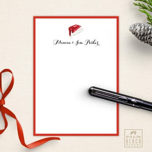 May include: A white stationery sheet with a red border, featuring a watercolor Santa hat and the names "Monica & Jim Parker" in elegant script. A black pen rests on the page, with a red ribbon and pinecone in the background. The Palm Beach Papery logo is in the bottom right.