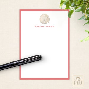 May include: A pink stationery notepad with a white seashell design and the name "Margaret Kendall" printed at the top. The notepad is on a beige textured background.