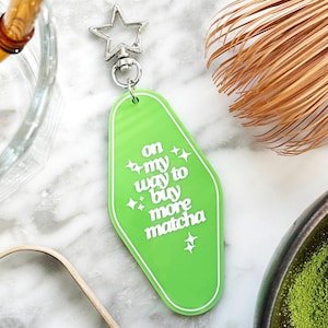 May include: A bright green keychain with the text "on my way to buy more matcha" in white. The keychain has a white border and a silver star-shaped clasp. The background includes matcha powder and a whisk.
