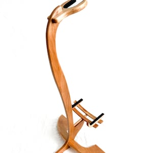 Puede incluir: Un soporte de guitarra de madera hecho a mano con un diseño curvo y fluido. El soporte es de madera marrón claro y presenta apoyos acolchados negros para la guitarra. La base es ancha para mayor estabilidad, y la altura total es de aproximadamente 91 cm.