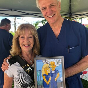 May include: A couple poses for a photo holding a framed cartoon portrait of themselves in a hospital operating room setting. The woman is wearing a white shirt with a black sash and a cross necklace. The man is wearing a blue scrub top. The cartoon portrait shows the couple in yellow scrubs with the man wearing a surgical mask.