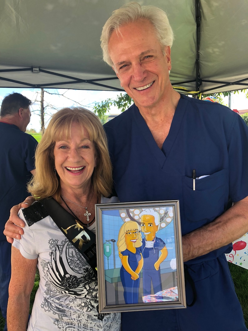 May include: A couple poses for a photo holding a framed cartoon portrait of themselves in a hospital operating room setting. The woman is wearing a white shirt with a black sash and a cross necklace. The man is wearing a blue scrub top. The cartoon portrait shows the couple in yellow scrubs with the man wearing a surgical mask.
