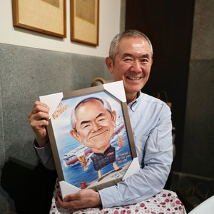 May include: A man is holding a framed caricature of a person with a beer in their hand. The caricature is in a blue and white frame with a white mat. The man is wearing a light blue button-down shirt and is smiling. The caricature has the text "Initial Service year 1992-2009" on it.