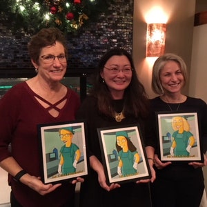 May include: Three people are holding framed cartoon portraits of themselves in scrubs. The portraits are in black frames and have a white background. The people are smiling and looking at the camera.