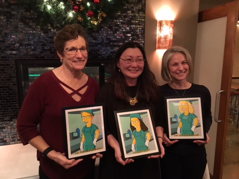 May include: Three people are holding framed cartoon portraits of themselves in scrubs. The portraits are in black frames and have a white background. The people are smiling and looking at the camera.