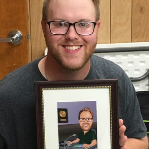 May include: A person holds a framed caricature of a person wearing glasses and a green shirt, sitting at a soundboard with a gold record on the wall behind them.