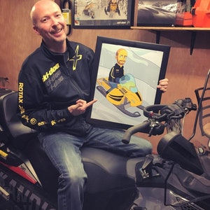 May include: A man wearing a black jacket with yellow accents and the text "Ski-Doo" is holding a framed cartoon illustration of a person riding a yellow snowmobile. The man is sitting on a gray snowmobile.
