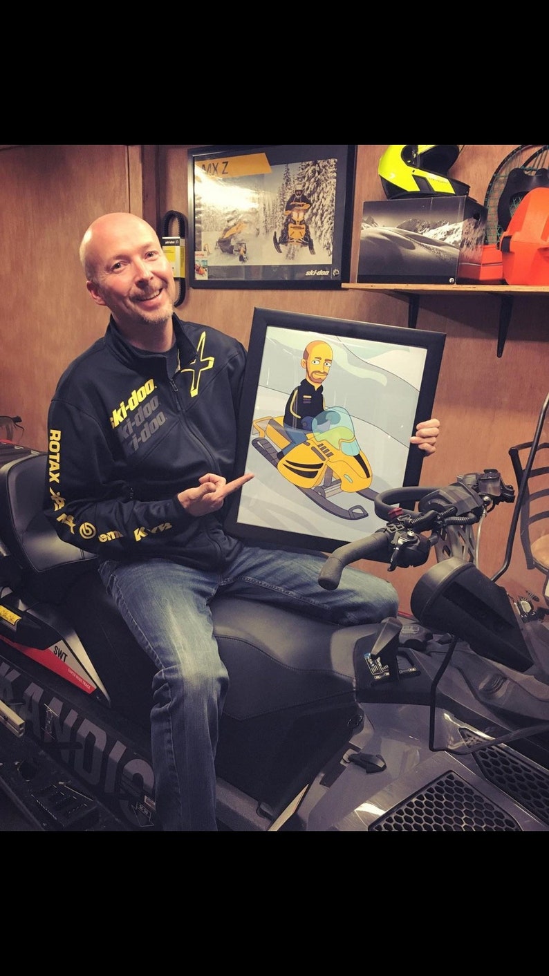 May include: A man wearing a black jacket with yellow accents and the text "Ski-Doo" is holding a framed cartoon illustration of a person riding a yellow snowmobile. The man is sitting on a gray snowmobile.