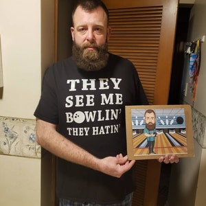 May include: A person wearing a black t-shirt with the text "They See Me Bowlin' They Hatin'" and a bowling ball graphic. The person is holding a framed cartoon illustration of a person bowling.