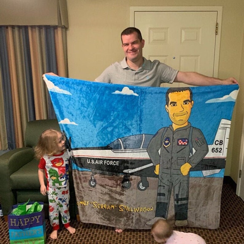 May include: A blue and gray blanket featuring a cartoon illustration of a pilot and a U.S. Air Force jet. The blanket has a blue sky background with white clouds. The text "U.S. AIR FORCE" is visible on the jet.