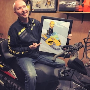 May include: A man wearing a black jacket with yellow accents and the text "Ski-Doo" is holding a framed cartoon illustration of a person riding a yellow snowmobile. The man is sitting on a gray snowmobile.