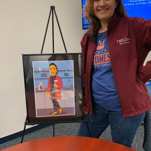 May include: A woman in a maroon jacket and blue shirt with the text "M CTO DNES CTCBeKind" stands in front of a framed illustration of a person in a red lab coat holding a beaker. The illustration is on an easel. A cupcake with white frosting is on a brown table.