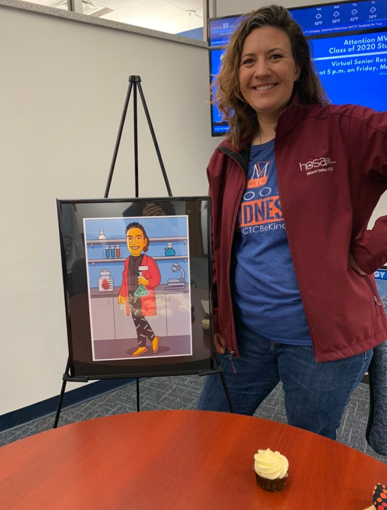 May include: A woman in a maroon jacket and blue shirt with the text "M CTO DNES CTCBeKind" stands in front of a framed illustration of a person in a red lab coat holding a beaker. The illustration is on an easel. A cupcake with white frosting is on a brown table.