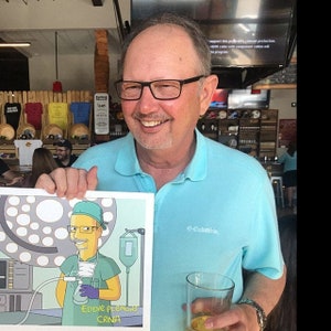 May include: A man holds a framed cartoon illustration of a person wearing a surgical mask and scrubs. The cartoon is titled "Eddie Plemmons, CRNA".